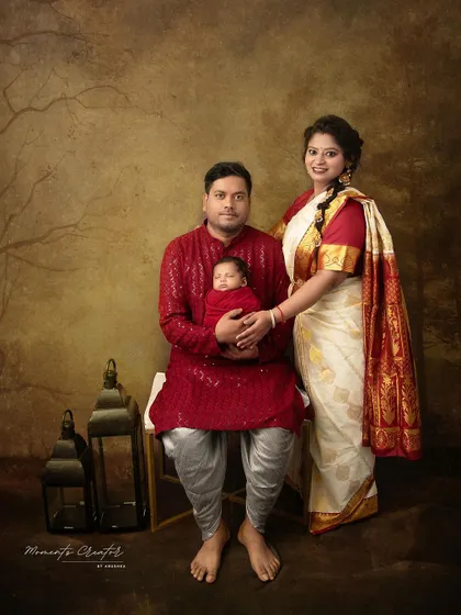 A full-length family portrait in traditional Indian clothing. The father sits holding his newborn while the mother stands beside them, creating a formal yet warm family heirloom.