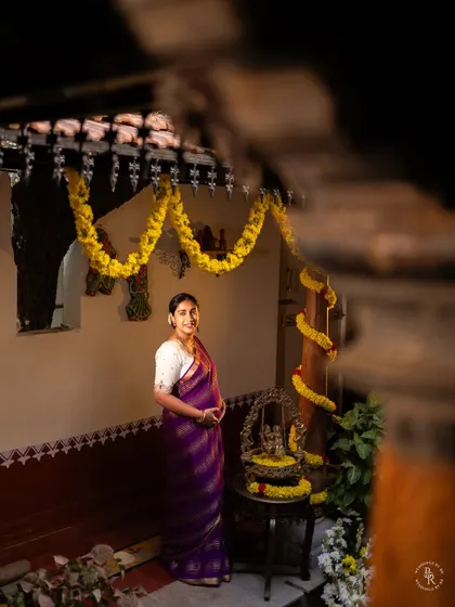 A full-length portrait of an expectant mother in a rich purple saree, standing proudly in a traditionally decorated home, embodying grace and happiness.
