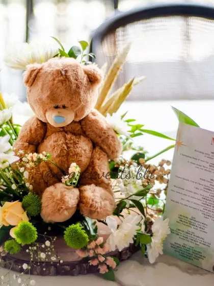 A charming centerpiece from a teddy bear-themed baby shower. A cute teddy bear sits amidst a bed of white and green flowers, next to a printed menu, showing attention to every little detail.