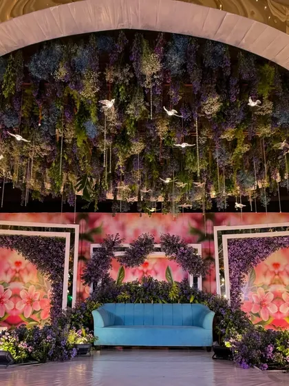 A magical reception stage with a ceiling of hanging florals and illuminated birds, set against a dreamy, floral-printed backdrop.