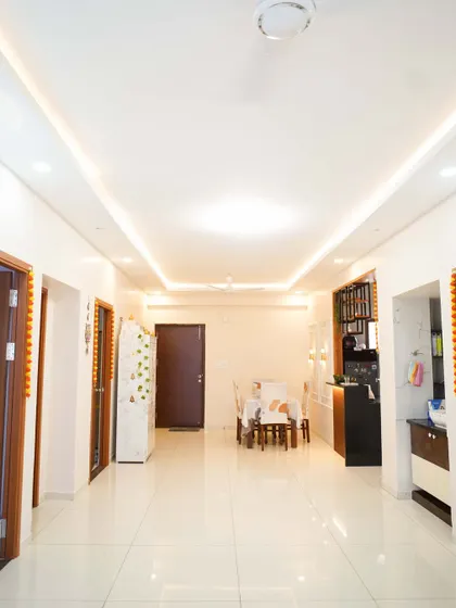A long shot of a spacious living area with clean white floors and a simple false ceiling with cove lighting, creating a bright and expansive feel.