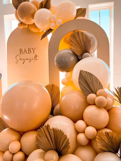 This modern baby shower setup features a Chiara wooden backdrop personalized with the baby's name. The neutral color palette with pastel balloons and dried palm leaves creates a chic and sophisticated look.