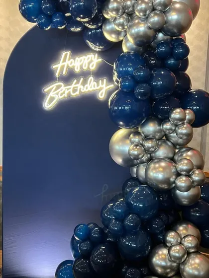 A close-up of a sophisticated balloon garland using premium chrome balloons in navy blue and silver, paired with a classic "Happy Birthday" neon sign.