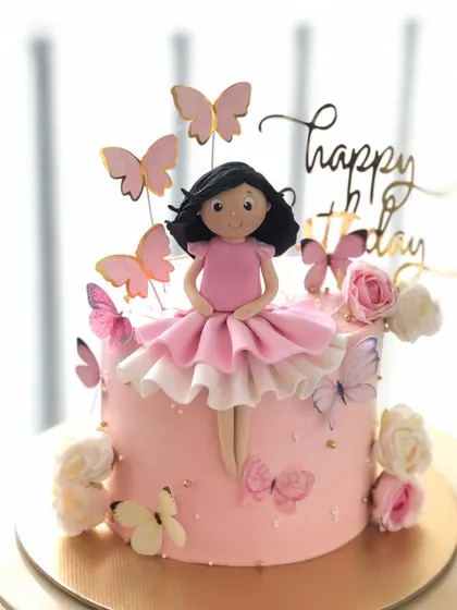 A delicate cake with a handcrafted doll topper, surrounded by pink butterflies and flowers.