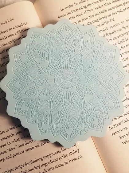 Our Mandala coaster in a calming icy blue color, placed inside a book. The intricate design feels both modern and timeless.