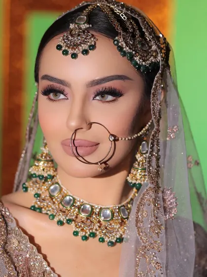 A close-up of a model wearing a full bridal look, including a green Polki choker, matching earrings, a stunning matha patti, and a classic nath.