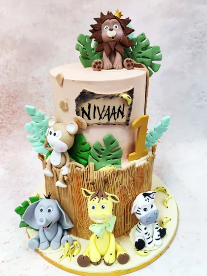 We create a wild celebration with this two-tiered jungle animal birthday cake. A crowned lion sits atop a tree-trunk tier, while a monkey, elephant, giraffe, and zebra gather below, all handcrafted for a memorable first birthday safari.
