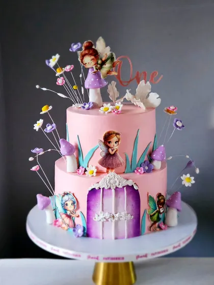 A dreamy, two-tiered fairyland cake for a first birthday. This design features a fairy house door, edible mushrooms, and a mix of handcrafted and printed fairies, all in a soft pink and purple color scheme.