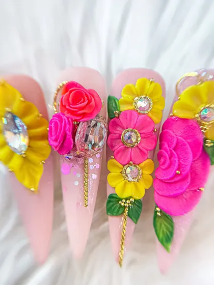 A stunning set of stiletto nails featuring elaborate 3D and 5D floral arrangements in bright pink and yellow, accented with large crystals and gold chains.