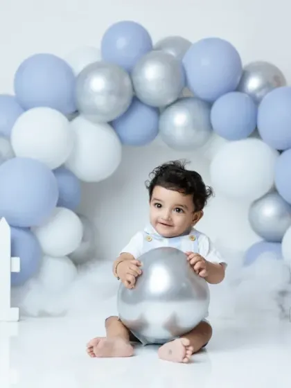 A sweet portrait from a first birthday shoot, with the baby boy sitting under a beautiful arch of blue, white, and silver balloons.