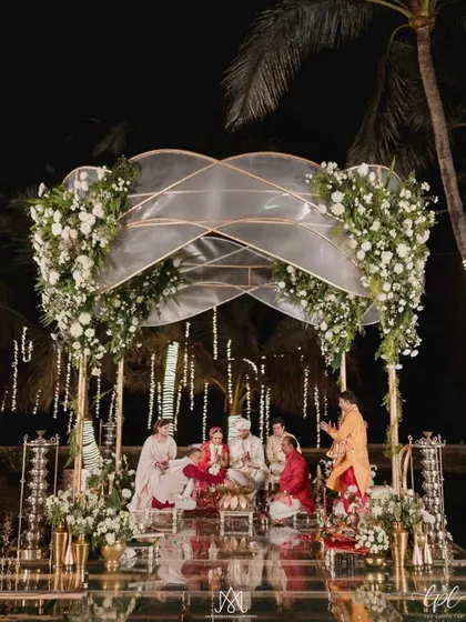 A couple's cherished moment during their wedding rituals, framed by a modern, elegant mandap with a translucent canopy and white floral arrangements, set over a pool.