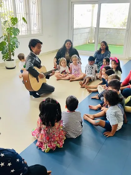 Children and educators sit together, completely engaged in a sing-along session with a live guitarist. Music helps build community and is a powerful tool for language development.