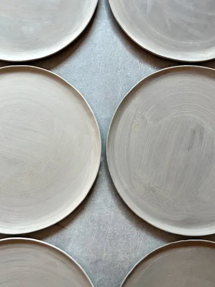 A symmetrical arrangement of unfired dinner plates, coated in a layer of slip. This shows the preparation stage before glazing, highlighting the raw, earthy texture of the clay.
