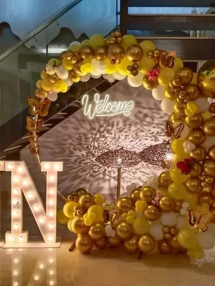 A welcome party setup with "NN" marquee letters and a yellow and gold crescent balloon arch.