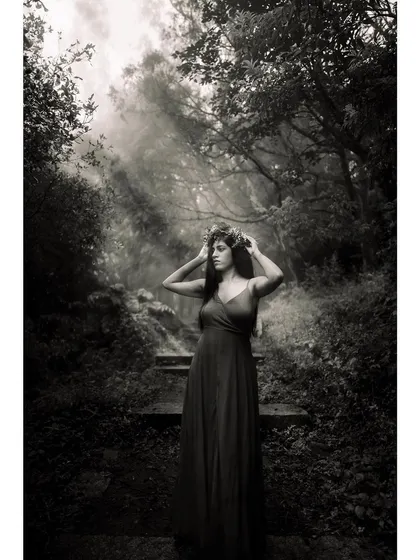 Black and white photo of a model in a dark dress and flower crown, standing in a misty forest with light rays