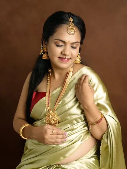 A close-up portrait of a beautiful mom-to-be adorned in traditional gold jewelry and a silk saree, looking down with a gentle smile.