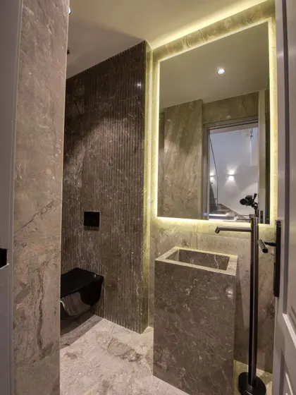 This powder room features a monolithic stone pedestal sink and a floor-mounted faucet. The back-lit mirror and fluted stone accent wall create a sophisticated, moody atmosphere.