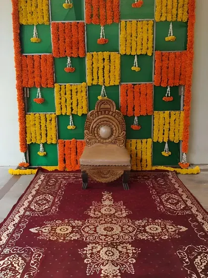 A classic floral backdrop using a checkerboard pattern of orange and yellow marigold garlands against a green base. This type of versatile stage decoration is suitable for various traditional Indian ceremonies.