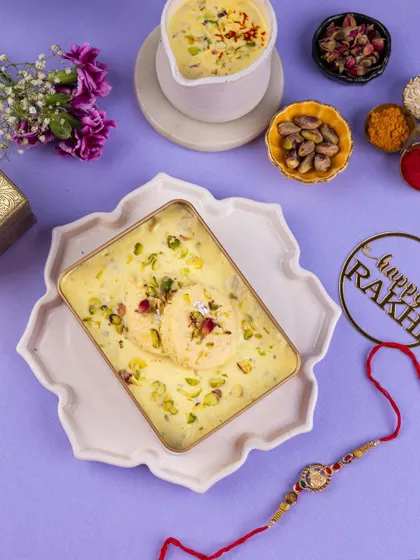 A top-down view of a Rasmalai dessert for Raksha Bandhan, styled with a rakhi and pistachios on a calming lavender surface.
