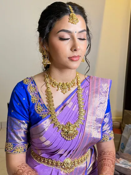 A beautiful South Indian bridal look featuring a soft, messy bun and traditional gold jewellery. The makeup is kept fresh and glowing to match the lavender silk saree.
