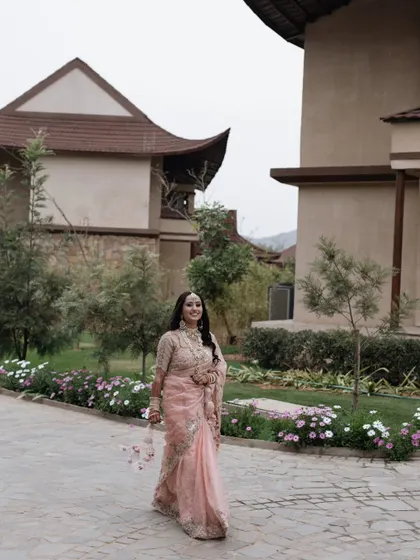 The bride walks along a garden path, her beautiful pink saree and joyful expression capturing the essence of a daytime wedding event.