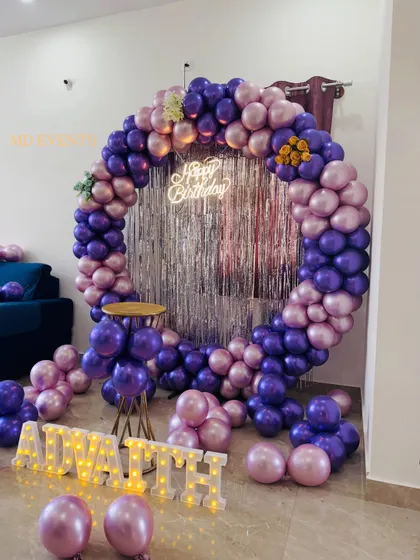 I love working with bold color palettes. This birthday decoration uses a circular frame wrapped in metallic purple and pink balloons, set against a silver fringe curtain for extra sparkle. The setup is completed with marquee letters for a personalized touch.