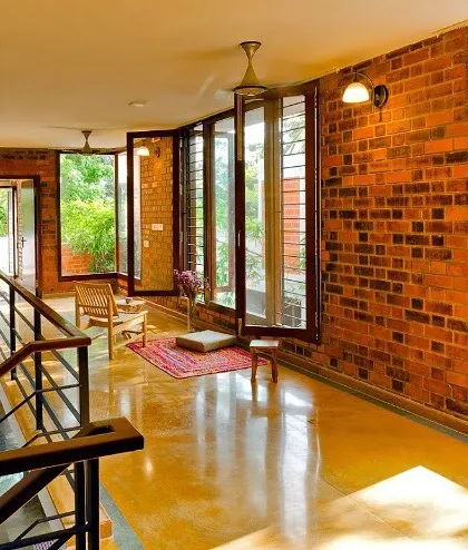 This corridor in the Vidyut Kapoor Residence is lined with floor-to-ceiling windows on one side and an exposed brick wall on the other. The design maximizes natural light and offers continuous views of the garden, blurring the line between inside and out.