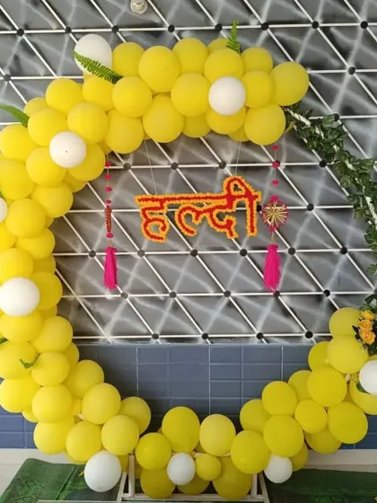 A modern take on Haldi decor, this circular backdrop is made of bright yellow and white balloons. A custom 'Haldi' sign in Hindi script hangs in the center, accented with green vines for a fresh look.
