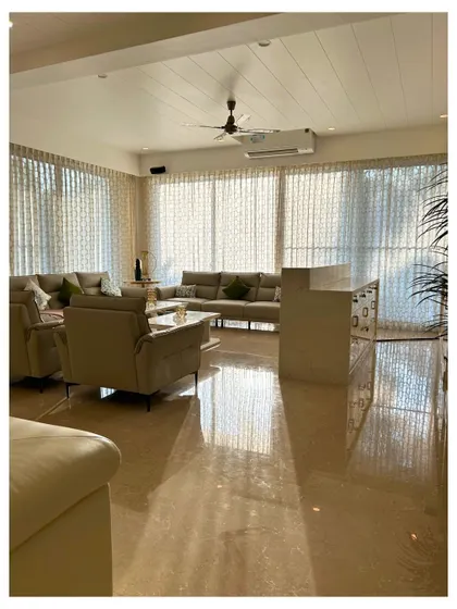 Sunlight streams into this bright living room, illuminating the polished marble floors and highlighting the elegant arrangement of neutral-toned furniture and sheer, flowing curtains.