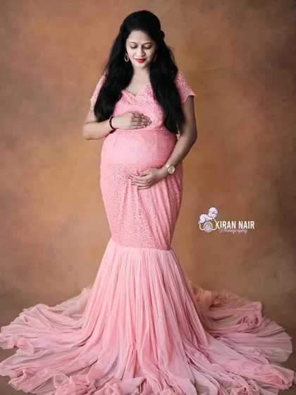 A full-length portrait in a pink lace mermaid gown with a flowing train, set against a warm, neutral studio backdrop.