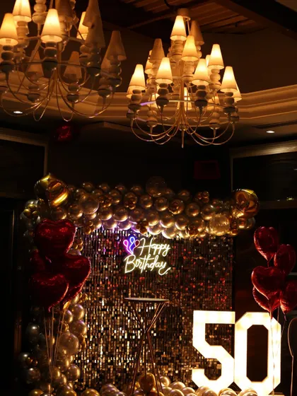 The decor for a 50th milestone celebration, featuring a shimmering gold backdrop, heart-shaped balloons, and large illuminated numbers.