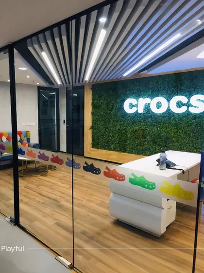 A view into the Crocs office reception from the outside, highlighting the playful branding elements. A colorful decal of Crocs shoes on the glass partition and a living green wall behind the logo create a fun and memorable entrance.