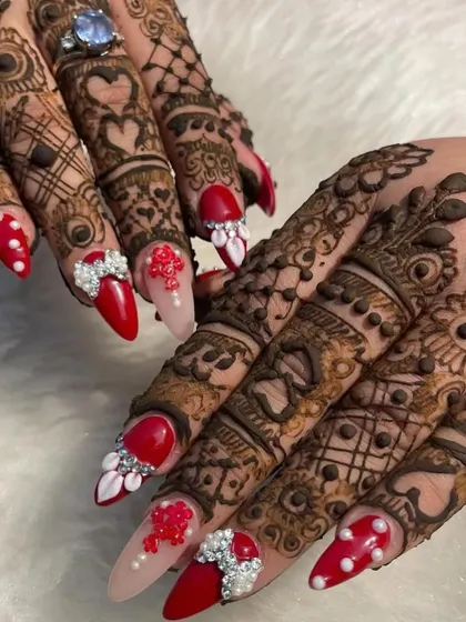A bold and beautiful bridal nail design. This set features vibrant red nails with pearl dots, intricate 3D floral and bow embellishments, and sparkling rhinestone accents, all designed to stand out alongside the bridal mehendi.