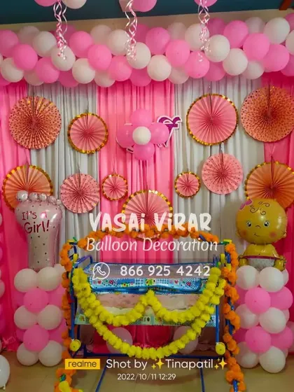 This naming ceremony setup for a baby girl uses a classic pink and white color scheme. The backdrop is made of drapes and paper fans, framed by a full balloon arch, with a decorated cradle ready for the baby.
