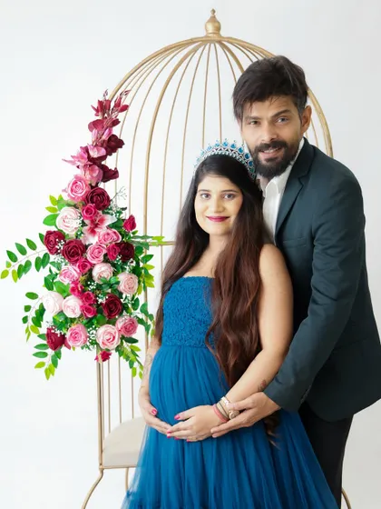 A stylish couple's portrait featuring a beautiful birdcage prop adorned with flowers. The mom-to-be looks regal in her blue gown and tiara.