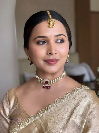 A close-up portrait of a bride with a very clean and minimalist makeup look. The focus is on perfect skin, subtly defined eyes, and a natural lip color, paired with a simple gold saree.