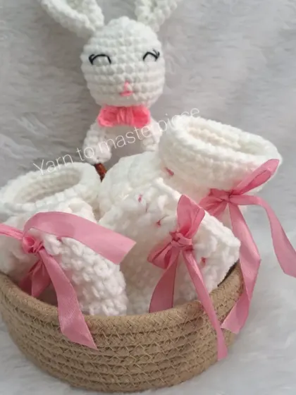 This newborn gift hamper includes soft white booties with pink ribbons and a matching bunny rattle, all presented in a charming woven basket.