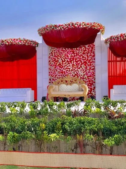A daytime view of a spectacular outdoor wedding stage. The vibrant red floral arrangements and drapery stand out beautifully against the open sky, creating a powerful and photogenic setting.