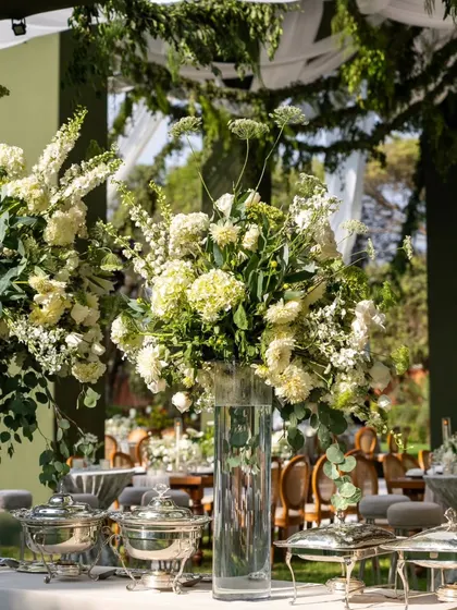 A large, lush arrangement of white and green florals in a clear glass vase, serving as a centerpiece for a buffet table. The arrangement adds height and drama to the food display.