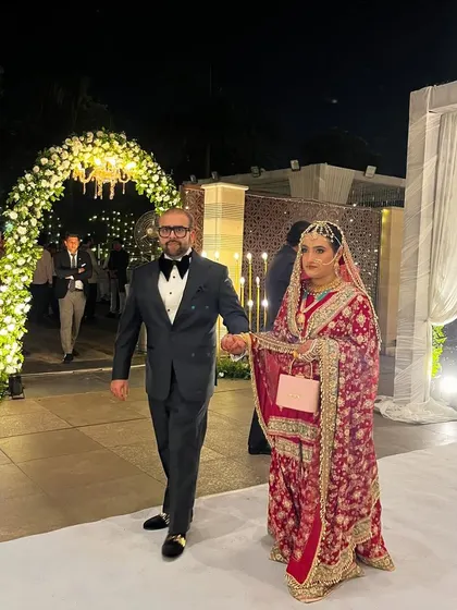 The Walima bride and groom make a grand entrance. They walk down a pristine white aisle, framed by elegant floral arches and warm lighting, creating a sophisticated and timeless moment.