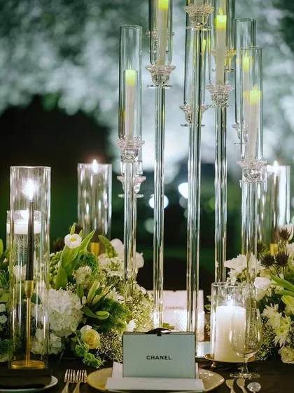 The soft glow of candles illuminates the intricate details of the Chanel-themed tablescape. This image captures the balance of modern glass candelabras and classic white floral arrangements, creating a look of effortless sophistication.