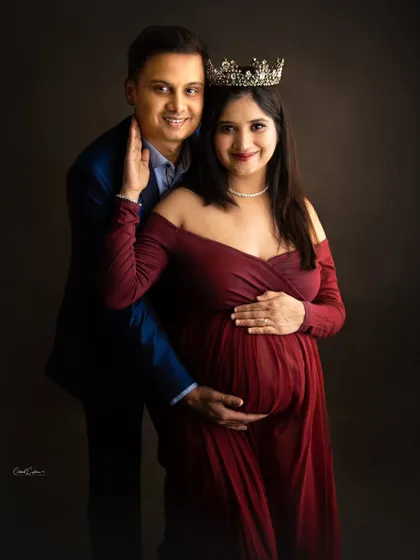 A royal-themed couple's portrait. The mom-to-be wears a crown as her partner stands lovingly behind her, both sharing a smile.