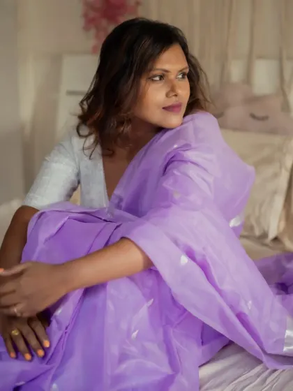 A relaxed indoor portrait on a bed. The model, wrapped in a beautiful lavender saree, looks away thoughtfully, creating a candid and personal lifestyle image.