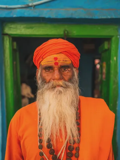 A close-up portrait of a sadhu in Varanasi. His gaze is direct, and his face, marked with tilak, tells a story of devotion and resilience against the vibrant blue and green of a local doorway.