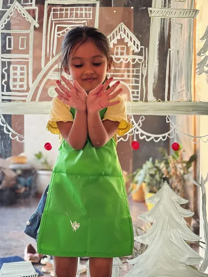 A candid shot of a little girl with paint on her hands, standing in front of a window painting. Art can be messy, and that's all part of the fun.