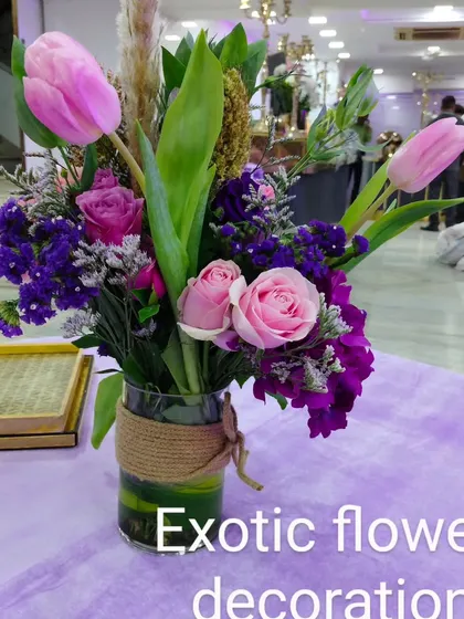 A modern table centerpiece for an event, featuring pink tulips, roses, and purple statice in a glass vase wrapped with jute rope. This style is perfect for corporate events or contemporary celebrations.