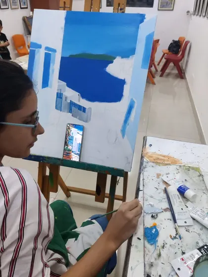 Using a phone for reference, the student carefully adds details to the architecture in her acrylic painting, ensuring accuracy in her depiction.