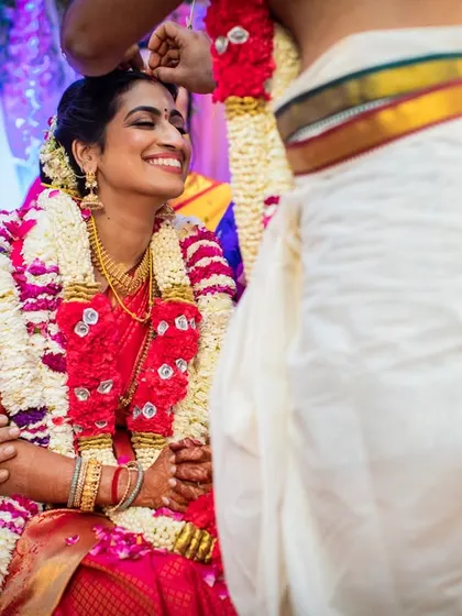 The bride's radiant smile as the groom performs a ritual, with her father looking on proudly. A beautiful multi-generational moment.