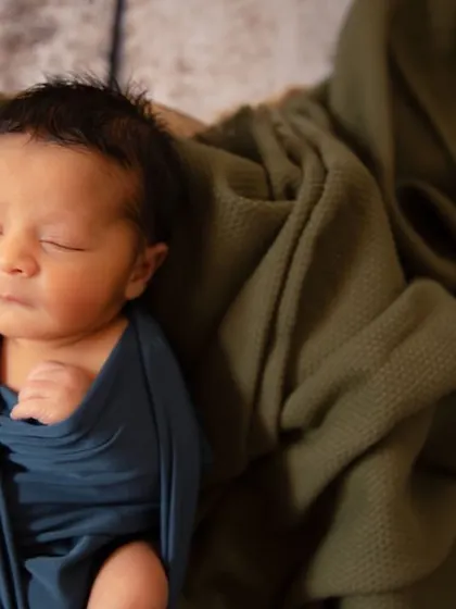 A beginning of all things. A newborn wrapped in a deep blue swaddle rests against an olive green blanket, creating a beautiful, earthy color combination.