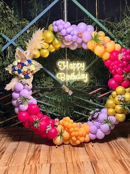 A vibrant and colorful circular balloon wreath. This decoration is perfect as a photo backdrop and incorporates multi-colored balloons, pampas grass, and floral elements.